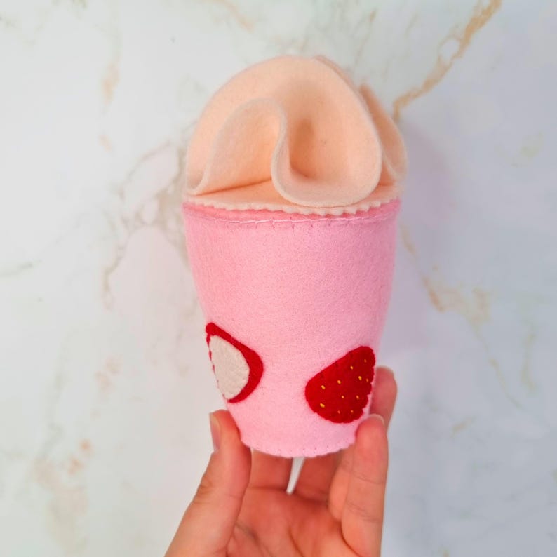 Puede incluir: Un batido de fresa de fieltro rosa con una cobertura de crema batida de fieltro rosa. El batido tiene dos rodajas de fresa en el lateral.