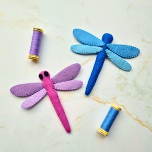 Könnte beinhalten: Zwei Libellen-Ornamente aus Filz, eine in Rosa- und Violetttönen und eine in Blautönen, begleitet von zwei Nähgarnrollen, eine violette und eine blaue, jeweils mit einer Längenangabe von 100m oder 110yds beschriftet.