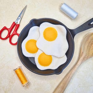 May include: Three felt fried eggs with yellow yolks in a black cast iron skillet. A wooden spoon, red scissors, and spools of thread in white and yellow are arranged around the skillet on a light-colored surface.