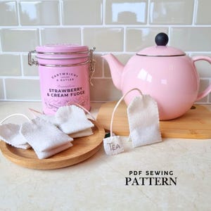 Può includere: Una teiera rosa con un pomello nero è appoggiata su un tagliere di legno accanto a una scatola rosa di Strawberry & Cream Fudge. Diverse bustine di tè bianche sono su un piatto di legno e una ha un'etichetta con la scritta "Tea". Il testo "PDF SEWING PATTERN" è in basso.
