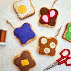May include: A collection of felt toast ornaments featuring various toppings including a fried egg, strawberry slices, purple jam, banana slices, and a butter square, accompanied by a spool of brown thread and red-handled metal scissors.