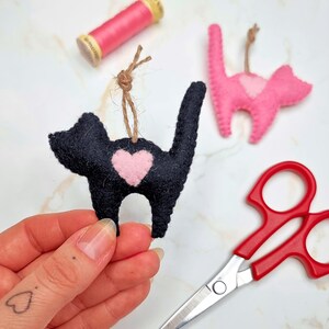 May include: Black felt cat ornament with a pink heart, held by a hand. A pink cat ornament and a spool of pink thread are in the background. Red scissors are in the lower right corner. The ornaments have twine hangers.