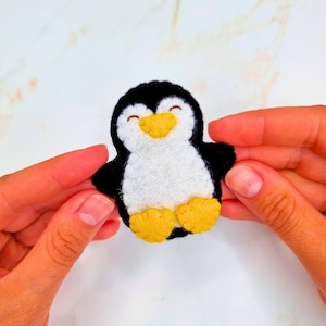May include: A small felt penguin applique featuring a black body, a white belly and face, a yellow beak, and two yellow feet. The penguin has a simple stitched expression with closed eyes and a small smile, crafted from soft felt material.