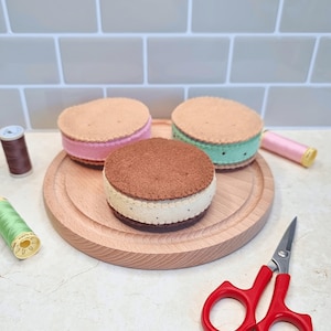 May include: Three felt ice cream sandwiches in brown, pink, and green, sit on a wooden cutting board. The sandwiches have stitched edges and are surrounded by sewing supplies, including thread spools and red scissors.