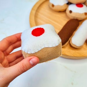 Puede incluir: Un pastel de juguete de fieltro con glaseado blanco y una cereza roja en la parte superior. El pastel se está sosteniendo en una mano.