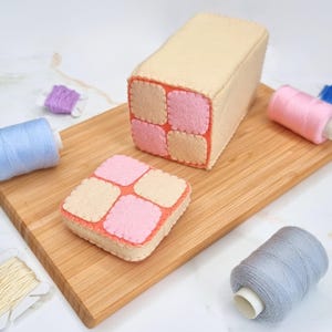 May include: A felt play food set featuring a loaf of bread and a slice of bread. The bread is beige with pink and beige squares. The bread is on a wooden chopping board.