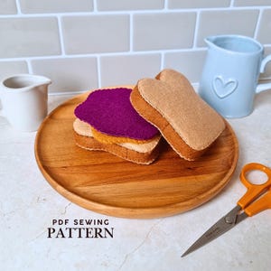 May include: A felt sandwich toy consisting of two tan bread slices, a yellow cheese layer, and a purple jelly layer, resting on a round wooden plate. The text PDF SEWING PATTERN is visible in the lower left corner, alongside orange handled scissors.