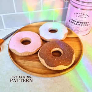May include: Three handmade felt donuts in pink, white, and brown, each featuring small stitched sprinkles. The donuts are arranged on a round wooden plate next to a pair of metal scissors and a pink tin labeled Cartwright & Butler Strawberry Cream Fudge.