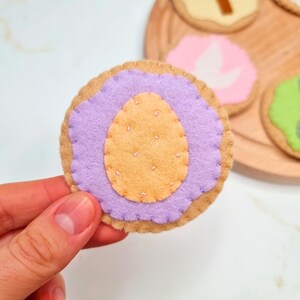 May include: A felt Easter cookie with purple icing and a yellow egg in the centre. The cookie is decorated with white stitching.