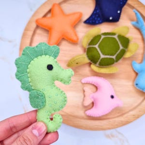 Puede incluir: Criaturas marinas de fieltro hechas a mano, incluyendo un caballito de mar verde, una estrella de mar naranja y una tortuga verde. Otras criaturas incluyen un pez rosa y una raya azul. Un caballito de mar es sostenido por una mano.