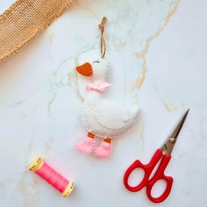 May include: A handmade white felt goose ornament with an orange beak, pink bow, and pink booties. The goose is suspended by a brown string. A spool of pink thread and red scissors are also in the image.