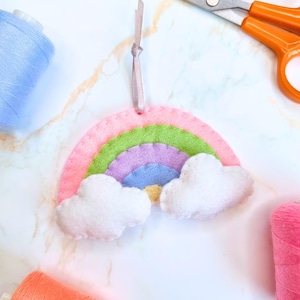 May include: A felt rainbow decoration with pink, green, purple, and blue arches, and two white cloud accents. A pink ribbon is attached for hanging. Spools of blue and pink thread and orange scissors are also visible.