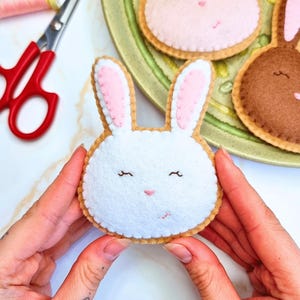 May include: A white felt bunny with pink inner ears, a light brown outline, and embroidered facial features. Additional felt bunnies in pink and brown are on a green plate. Scissors and thread are in the background.