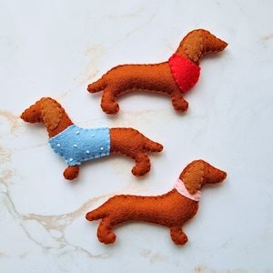 May include: Three felt dachshund dog ornaments in shades of brown, with tan ears and snouts. One dog wears a red scarf, another a blue sweater with white dots, and the third a pink scarf. The dogs are arranged on a white marble surface.