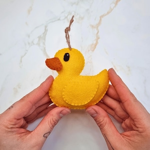 Puede incluir: Adorno de pato de fieltro amarillo hecho a mano con pico marrón y ojo negro. El pato tiene una cuerda marrón para colgar. El adorno se sostiene entre dos manos.
