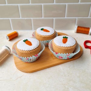 Puede incluir: Tres decoraciones de pastel de zanahoria de fieltro sobre una tabla de madera. Cada pastel tiene una parte superior blanca con un diseño de zanahoria de fieltro y un borde festoneado. Los pasteles están en moldes de papel decorativos. Bobinas de hilo naranja y tijeras rojas en el fondo.