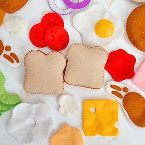 Peut inclure: Un ensemble d'aliments factices en feutrine comprenant deux tranches de pain beige, un œuf au plat, des tranches de tomate rouge, une tranche de fromage jaune avec un trou, des champignons blancs, des tranches d'avocat vert et des morceaux de viande brune.