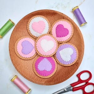 May include: Six felt coasters with heart designs in pink, purple, and white sit on a wooden plate. The coasters have a scalloped edge. Spools of thread in green, pink, and purple, and red-handled scissors are also visible.