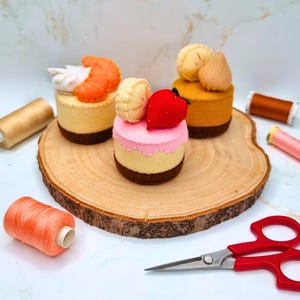 Può includere: Tre torte in feltro con vari condimenti, tra cui un gambero, una fragola e panna. Le torte sono su una fetta di legno, con rocchetti di filo e forbici nelle vicinanze. Le torte hanno una base marrone, un centro giallo chiaro e una parte superiore rosa.