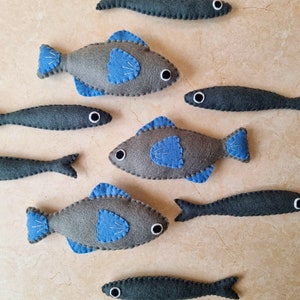 May include: A collection of felt fish toys in various shades of gray with blue accents. The fish have different sizes and shapes, with some having a more realistic appearance and others being more stylized.