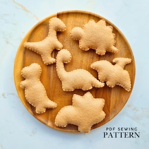 May include: Six tan felt dinosaur shapes arranged on a round wooden plate. The plush figures include various species with visible stitched edges. The text PDF SEWING PATTERN is printed in black in the bottom right corner of the image.