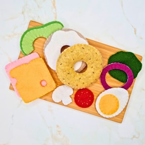 Puede incluir: Conjunto de comida de fieltro para juegos de simulación, que incluye un bagel, queso, aguacate, huevo, champiñón, tomate y cebolla. Las piezas de comida están dispuestas en una tabla de cortar de madera.