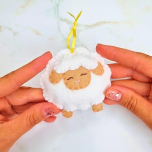 Puede incluir: Un adorno de fieltro con forma de oveja en color blanco y tostado, con bordes festoneados, cuerpo de textura suave y una pequeña nariz bordada en rosa. La oveja tiene los ojos cerrados y una cinta amarilla brillante para colgarla.