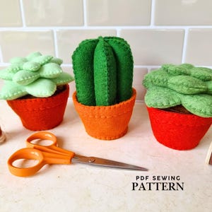 May include: Three felt succulent and cactus plants in orange pots. The cactus is green and the succulents are light green. A pair of orange-handled scissors lies in front of the plants. The text "PDF SEWING PATTERN" is at the bottom.