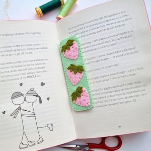 May include: A rectangular light green felt bookmark featuring three embroidered pink strawberries with dark green leaves, placed on an open book page with printed text and a line drawing of two figures kissing, near two spools of thread and scissors.