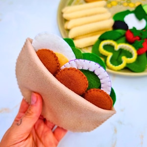 Puede incluir: Un sándwich de pita de fieltro con falafel marrón, cebolla blanca, lechuga y otros rellenos. Un plato con patatas fritas y ensalada de fieltro está en el fondo. El sándwich está sostenido por una mano.