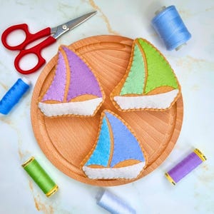 May include: Three colourful felt sailboat ornaments in purple, green, and blue, each with a white base and tan hull, arranged on a wooden tray. Red scissors and spools of thread in various colours, including blue, green, and purple, are scattered around the tray.