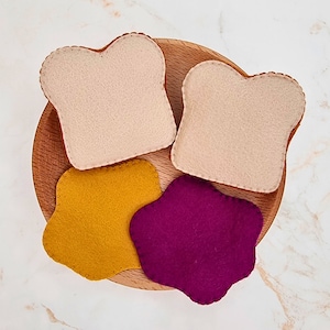 May include: Two beige felt bread slices alongside one mustard yellow and one deep purple felt shape with stitched edges. These four flat felt pieces are arranged on a round wooden plate, showcasing a soft texture and simple, clean outlines.