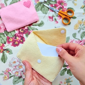 May include: A pale yellow felt envelope with a white snap closure containing a white card. A separate pink felt rectangle with a small pink heart appliqué and orange-handled scissors are positioned nearby on a floral patterned fabric surface.