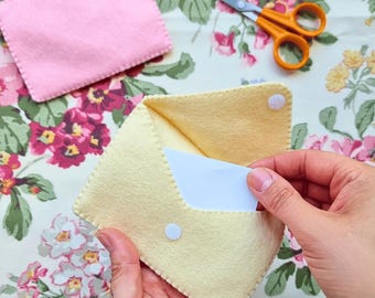 May include: A pale yellow felt envelope with a white snap closure containing a white card. A separate pink felt rectangle with a small pink heart appliqué and orange-handled scissors are positioned nearby on a floral patterned fabric surface.