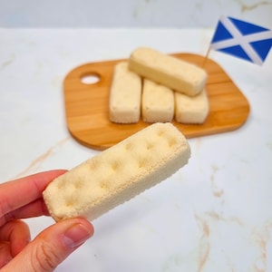 Puede incluir: Primer plano de una mano sosteniendo una galleta corta de fieltro marrón claro. La galleta tiene una superficie texturizada y está sobre un fondo blanco. Hay cuatro galletas más en una tabla de cortar de madera en el fondo.