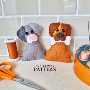 Pode incluir: Dois ornamentos de feltro em forma de cão, um cinzento e um castanho, com detalhes costurados e um osso branco. Ao lado, encontram-se uma bobine de fio laranja, tesouras com cabos laranja e uma almofada de alfinetes. Texto: PDF SEWING PATTERN.