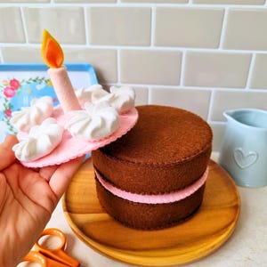 May include: A felt toy cake consisting of two brown circular layers with a pink filling layer. A separate pink circular top piece features white felt dollops of frosting and a single upright candle with a yellow and orange flame.