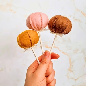 Puede incluir: Tres piruletas con forma de calabaza de fieltro en tonos rosa, naranja y marrón. Cada piruleta está unida a un palito blanco con un pequeño lazo blanco. Las calabazas tienen detalles cosidos.