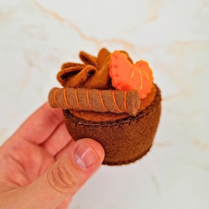 May include: A handmade felt cupcake in shades of brown and orange. The cupcake features a swirl of brown frosting, a small orange felt strawberry, and a brown wafer cookie. The cupcake is held in a hand.