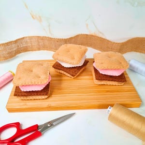 Może przedstawiać: Trzy filcowe s'mores na drewnianej desce. Każdy s'more ma beżowy krakers, brązową warstwę czekolady i różową warstwę pianki marshmallow. Czerwone nożyczki i szpulki nici w tle.