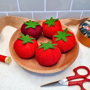 May include: A wooden plate holds five handmade felt tomatoes. The tomatoes are primarily red with green felt leaves. One tomato is a darker shade of red. Sewing supplies, including red scissors and thread, are also visible.