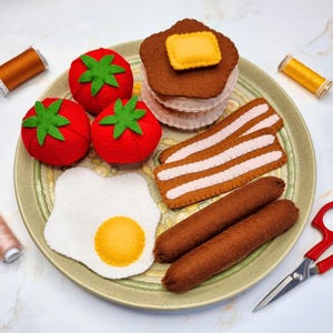 May include: A felt breakfast set on a green plate, including three red tomatoes with green tops, a stack of pancakes with a yellow butter square, bacon strips, two sausages, and a fried egg. Sewing supplies are in the background.