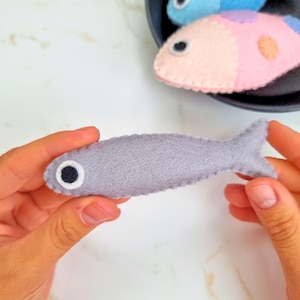May include: A handmade gray felt fish with a black and white eye. The fish is held in a person's hands. Other colorful felt fish are in the background, including pink and blue fish.