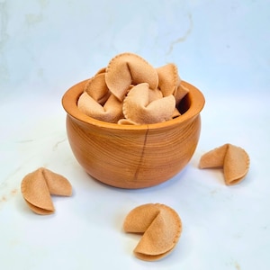 Puede incluir: Un cuenco de madera lleno de galletas de la fortuna de color beige. Tres galletas de la fortuna están esparcidas alrededor del cuenco sobre una superficie blanca y azul. El cuenco es de madera y tiene forma redonda.