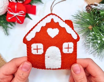 May include: A felt gingerbread house ornament in a deep orange-red color with white felt accents representing snow on the roof, a central heart shape, two windows with cross-shaped detailing, and a rounded door, featuring a brown hanging string.