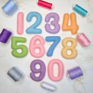 May include: A set of felt numbers from zero to nine in various pastel colors, including pink, blue, purple, orange, green, and yellow, arranged alongside several spools of matching sewing thread in shades of blue, pink, purple, and grey.