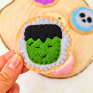 Op de afbeelding: Een rond viltornament met het gezicht van een groen Frankenstein-monster met zwart haar, gestikte ogen en een klein litteken. Het monster is gecentreerd op een lichtbruine ronde basis met een lichtblauwe geschulpte rand, gemaakt van zacht vilt.