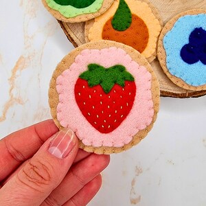 May include: A selection of handmade felt cookies, including a strawberry cookie with a red strawberry design on a pink centre. Other felt cookies include a blueberry, orange slice, and kiwi slice. The cookies are tan with a scalloped edge.