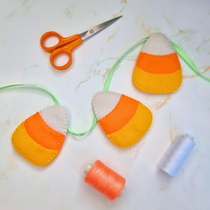 Op de afbeelding: Een vilt candy corn slinger met drie driehoekige stukken met witte, oranje en gele horizontale strepen bevestigd aan een lichtgroen lint. Oranje naaigaren en een klosje wit garen liggen in de buurt van een metalen schaar.