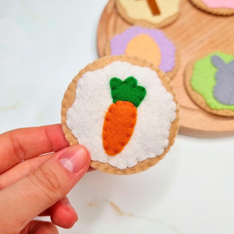 May include: A felt carrot cookie with a white frosting base and a green felt top. The cookie is held in a hand and is part of a set of felt cookies.